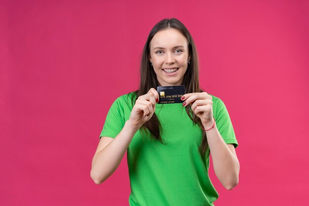 Giovane bella ragazza che indossa la maglietta verde che mostra la carta di credito con la faccia felice sorridente in piedi su sfondo rosa isolato