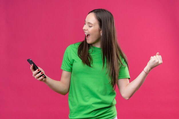 Giovane bella ragazza che indossa la maglietta verde che tiene smartphone uscito e felice alzando il pugno chiuso sorridendo allegramente gioendo il suo successo in piedi su sfondo rosa isolato