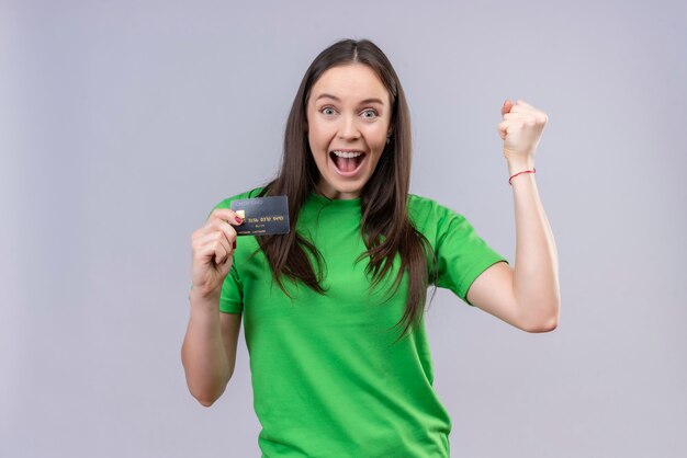 Giovane bella ragazza che indossa la maglietta verde è uscito e felice tenendo la carta di credito sorridendo allegramente alzando il pugno rallegrandosi del suo successo in piedi su sfondo bianco isolato