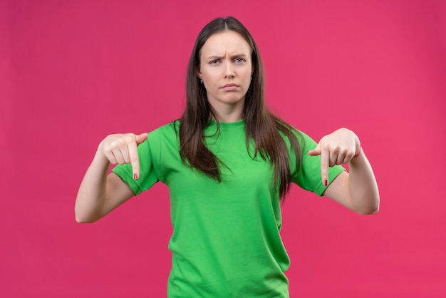 Giovane bella ragazza che indossa la t-shirt verde che guarda l'obbiettivo scontento con la faccia accigliata che punta con le dita verso il basso in piedi su sfondo rosa isolato