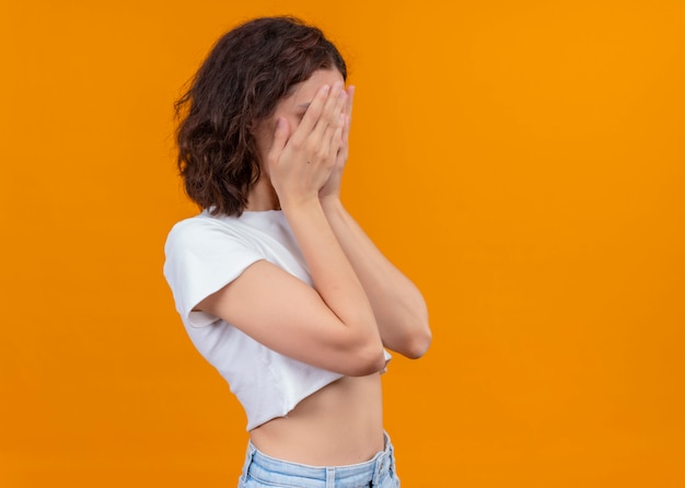 Giovane bella ragazza chiudendo il viso con le mani in piedi in vista di profilo su sfondo arancione isolato con spazio di copia