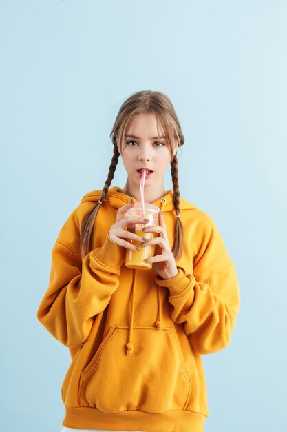 Giovane bella ragazza con due trecce in felpa con cappuccio che tiene in mano una tazza di plastica con succo d'arancia mentre guarda sognante a porte chiuse su sfondo blu