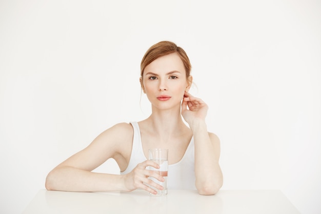 Giovane bella ragazza con pelle fresca pulita che giudica di vetro con acqua che si siede al tavolo. Stile di vita di salute e bellezza.