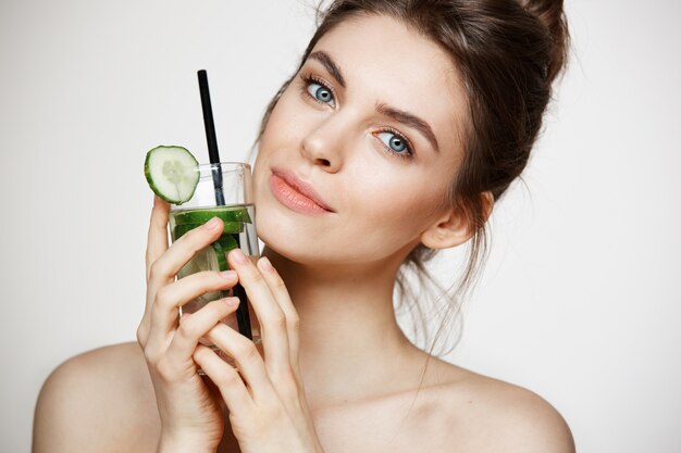 Giovane bella ragazza con pelle pulita perfetta che sorride esaminando macchina fotografica che tiene bicchiere d'acqua con le fette del cetriolo sopra fondo bianco. Nutrizione Sana.