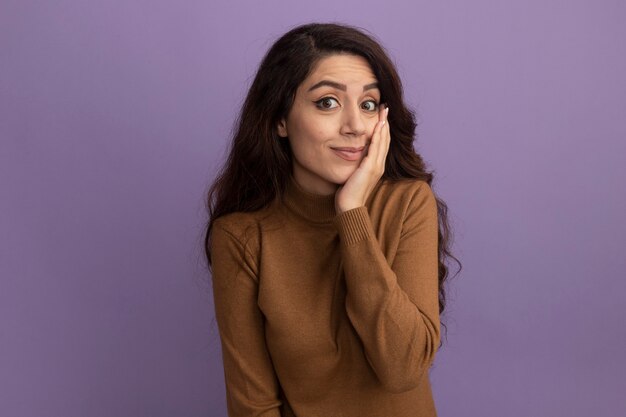 Giovane bella ragazza dispiaciuta che indossa maglione dolcevita marrone che mette la mano sulla guancia isolata sulla parete viola