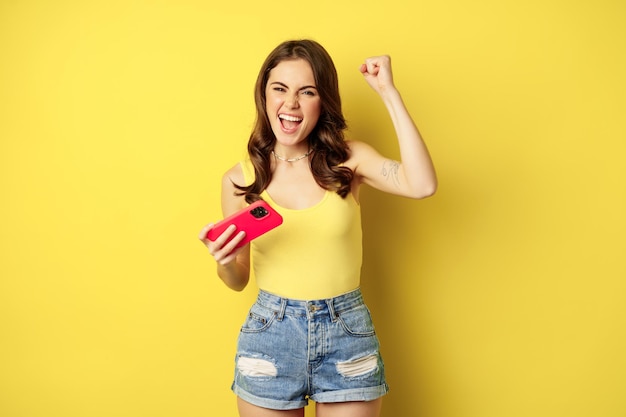 Giovane bella ragazza entusiasta che tiene lo smartphone orizzontalmente, gioendo, vincendo sul videogioco del telefono cellulare, in piedi su sfondo giallo.