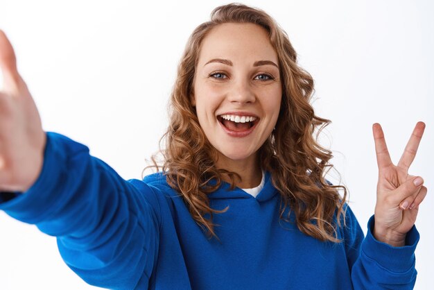 Giovane bella ragazza felice che prende selfie sul telefono cellulare allunga la mano e tiene lo smartphone che mostra la pace vsign e sorridente sfondo bianco