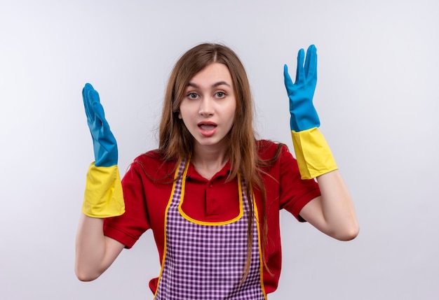 Giovane bella ragazza frustrata in grembiule e guanti di gomma che sembrano alzare le mani confuse