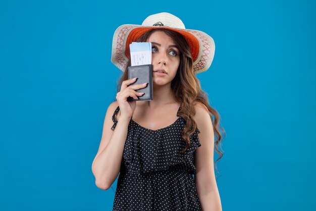 Giovane bella ragazza in abito a pois in cappello estivo in possesso di biglietti aerei cercando stanco e annoiato in piedi su sfondo blu