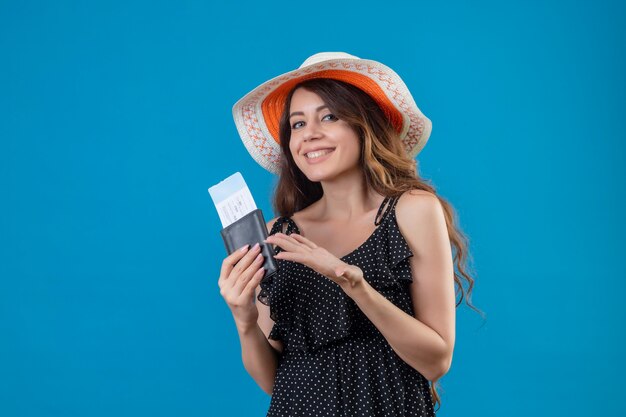 Giovane bella ragazza in abito a pois in cappello estivo in possesso di biglietti aerei che presentano con il braccio della sua mano sorridendo allegramente positivo e felice in piedi su sfondo blu