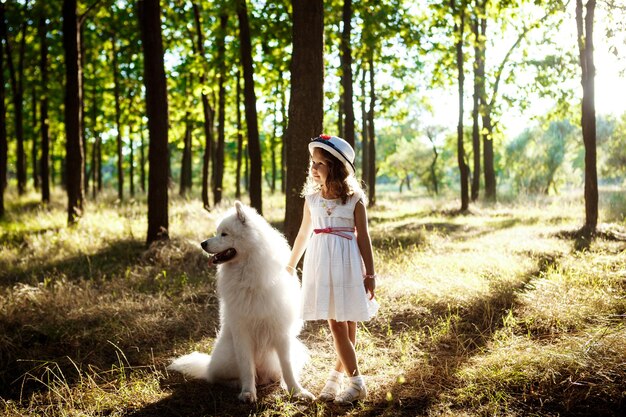Giovane bella ragazza in abito e cappello a piedi, giocando con il cane bianco nel parco al tramonto.