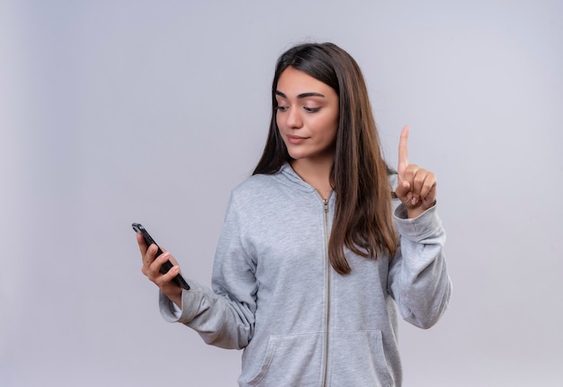 Giovane bella ragazza in felpa con cappuccio grigia che guarda al telefono con il sorriso sul viso che mostra il numero uno in piedi su sfondo bianco