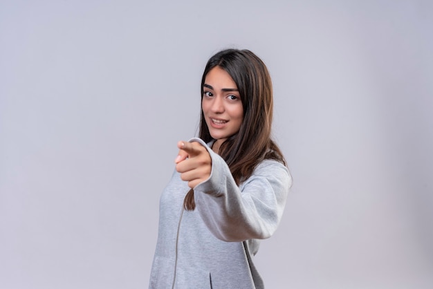 Giovane bella ragazza in felpa con cappuccio grigia che guarda l'obbiettivo con espressione insoddisfatta e che punta alla fotocamera in piedi su sfondo bianco