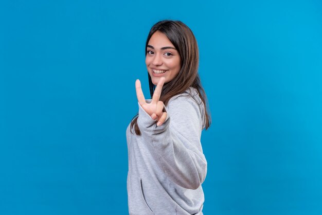 Giovane bella ragazza in felpa con cappuccio grigia che guarda l'obbiettivo con il sorriso sul viso che fa il gesto di pace in piedi su sfondo blu