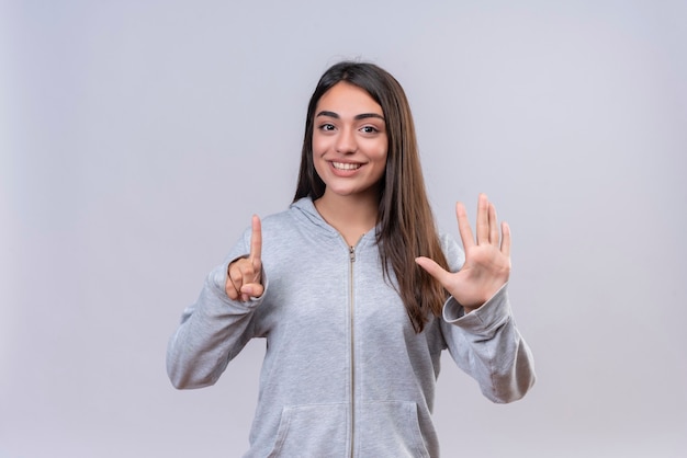 Giovane bella ragazza in felpa con cappuccio grigia che guarda l'obbiettivo con il sorriso sul viso che mostra uno e cinque numeri in piedi su sfondo bianco