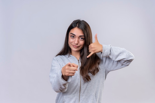 Giovane bella ragazza in felpa con cappuccio grigia che guarda l'obbiettivo con il sorriso sul viso che punta alla fotocamera che fa chiamarmi gesto in piedi su sfondo bianco