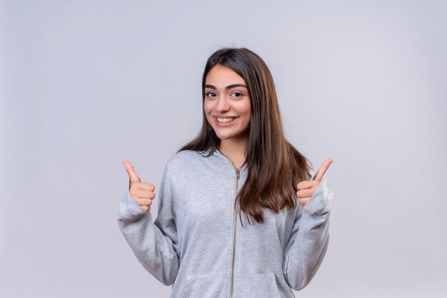 Giovane bella ragazza in felpa con cappuccio grigia che guarda l'obbiettivo con il sorriso sul viso facendo come gesto in piedi su sfondo bianco