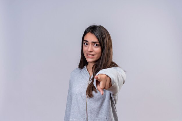 Giovane bella ragazza in felpa con cappuccio grigia che guarda l'obbiettivo con la faccia spaventata e che punta alla fotocamera in piedi su sfondo bianco