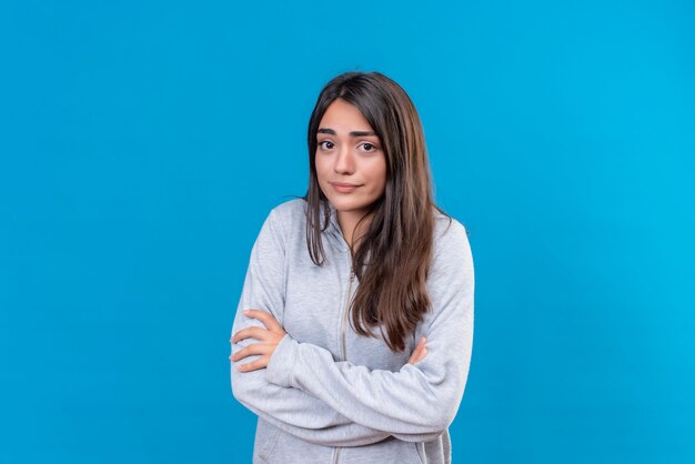 Giovane bella ragazza in felpa con cappuccio grigia che guarda l'obbiettivo con paura ansiosa emozione in piedi su sfondo blu