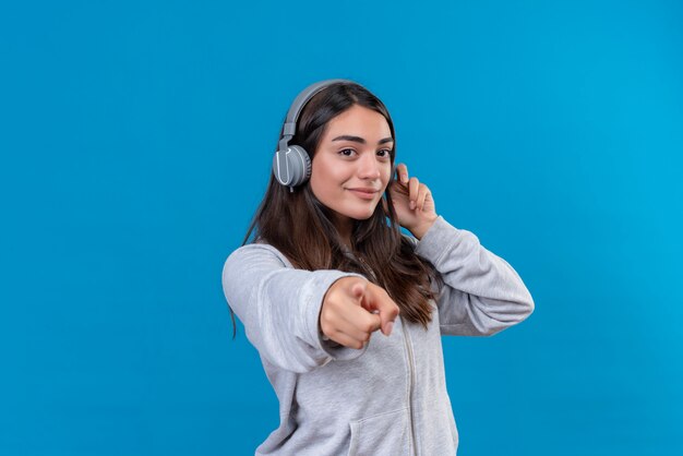 Giovane bella ragazza in felpa con cappuccio grigia che guarda l'obbiettivo con piacere e punta alla fotocamera in piedi su sfondo blu