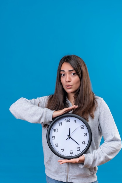 Giovane bella ragazza in felpa con cappuccio grigia che guarda l'obbiettivo con shock sul viso e tenendo l'orologio in piedi su sfondo blu