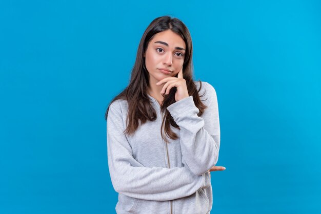 Giovane bella ragazza in felpa con cappuccio grigia che guarda l'obbiettivo con vista pensierosa e tenendo il dito sul viso in piedi su sfondo blu