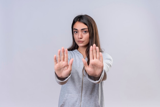 Giovane bella ragazza in felpa con cappuccio grigia che guarda l'obbiettivo con vista seriamente e facendo gesto di arresto in piedi su sfondo bianco