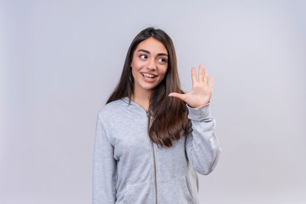 Giovane bella ragazza in felpa con cappuccio grigia che guarda lontano che mostra cinque con il sorriso sul viso in piedi su sfondo bianco