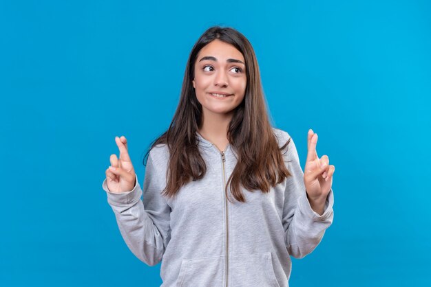 Giovane bella ragazza in felpa con cappuccio grigia che guarda lontano con emozione ansiosa che fa gesto di buona fortuna in piedi su sfondo blu