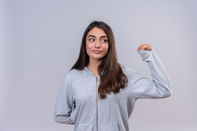 Giovane bella ragazza in felpa con cappuccio grigia che guarda lontano con il sorriso sul viso in piedi su sfondo bianco