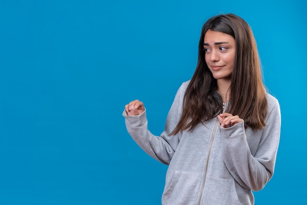 Giovane bella ragazza in felpa con cappuccio grigia che guarda lontano con la faccia spaventata in piedi su sfondo blu