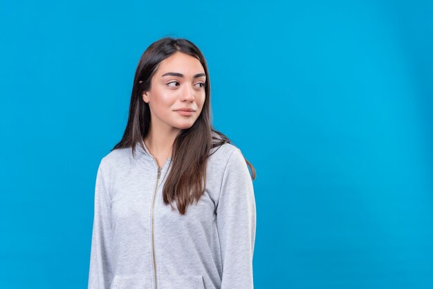 Giovane bella ragazza in felpa con cappuccio grigia che guarda lontano in piedi su sfondo blu