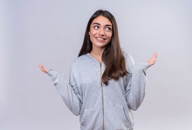 Giovane bella ragazza in felpa con cappuccio grigia che guarda lontano senza avere risposta, diffondendo le palme in piedi su sfondo bianco