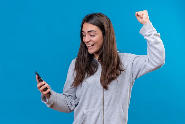 Giovane bella ragazza in felpa con cappuccio grigia che tiene telefono alla ricerca di telefono con emozione di piacere in piedi su sfondo blu