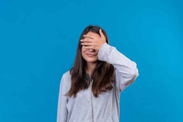 Giovane bella ragazza in felpa con cappuccio grigia con la mano sulla testa in piedi su sfondo blu