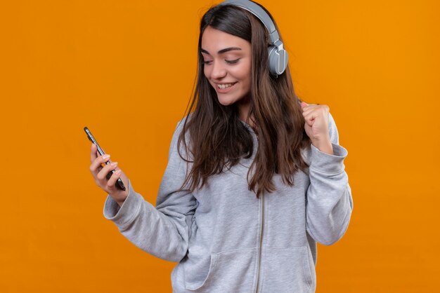 Giovane bella ragazza in felpa con cappuccio grigia con le cuffie guardando il telefono con il sorriso sul suo viso in piedi su sfondo arancione