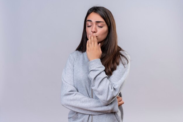 Giovane bella ragazza in felpa con cappuccio grigia con occhio chiuso baciare la mano in piedi su sfondo bianco