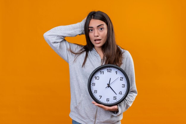 Giovane bella ragazza in felpa con cappuccio grigia guarda la fotocamera in piedi con la mano sulla testa per errore e l'altra mano con l'orologio sulla sua mano in piedi su sfondo arancione