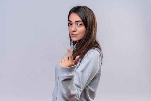 Giovane bella ragazza in felpa con cappuccio grigia guardando la telecamera fiducioso sul viso rivolto verso l'alto in piedi su sfondo bianco