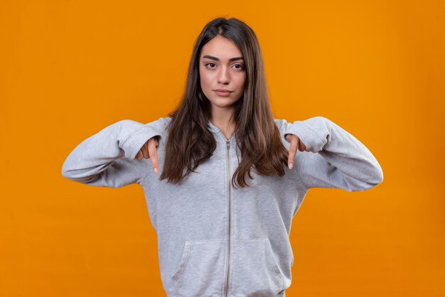 Giovane bella ragazza in felpa con cappuccio grigia rivolta verso il basso con dispiaciuto in piedi su sfondo arancione