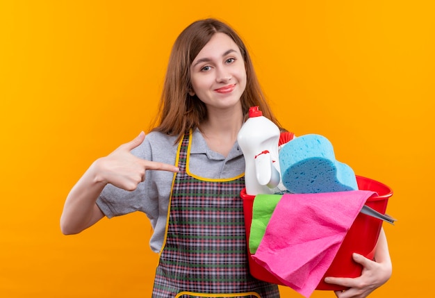 Giovane bella ragazza in grembiule che tiene secchio con strumenti di pulizia che punta con il dito indice ad esso sorridente fiducioso