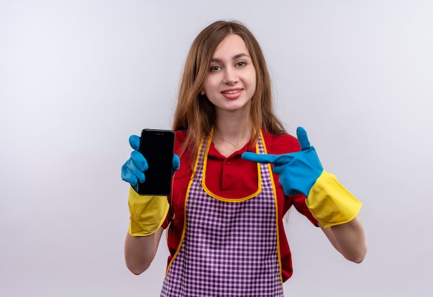 Giovane bella ragazza in grembiule e guanti di gomma che mostrano smartphone che punta con il dito sorridendo