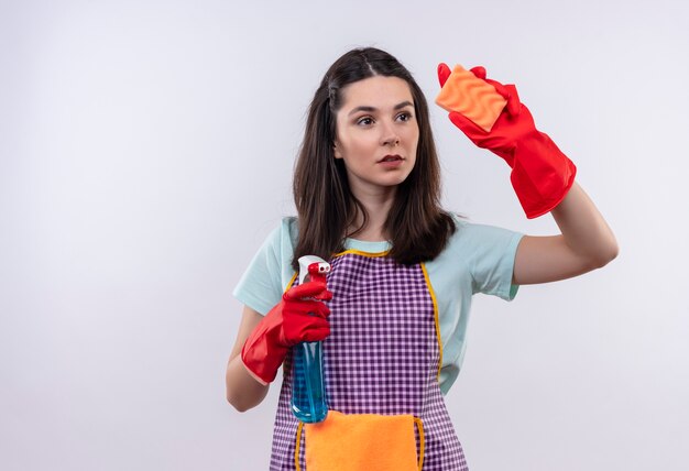 Giovane bella ragazza in grembiule e guanti di gomma che tengono spray per la pulizia e spugna andando a pulire guardando fiducioso