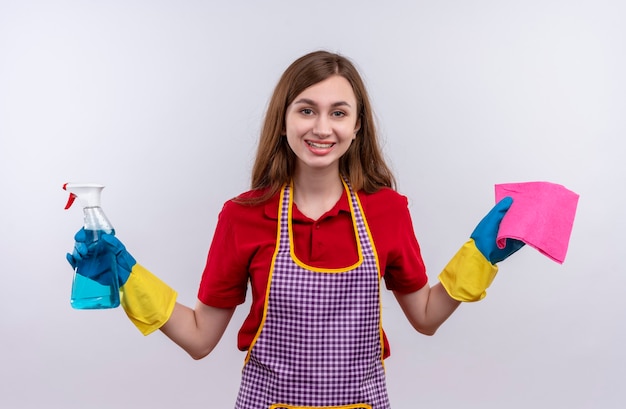 Giovane bella ragazza in grembiule e guanti di gomma che tengono spray per la pulizia e tappeto guardando la telecamera sorridendo allegramente, pronta per la pulizia su sfondo wjite