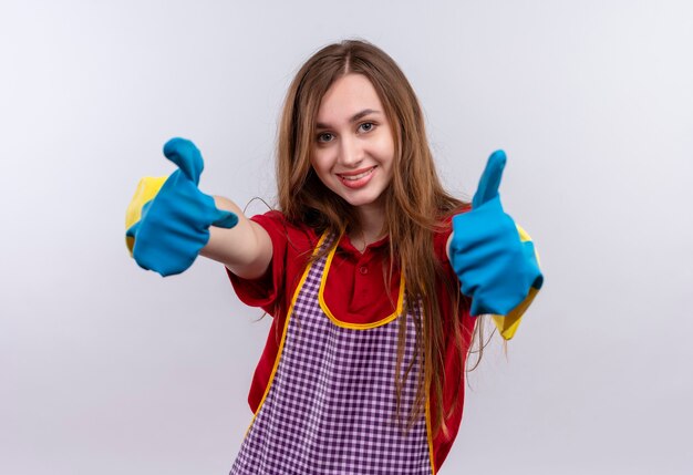 Giovane bella ragazza in grembiule e guanti di gomma sorridente che mostra i pollici in su con entrambe le mani