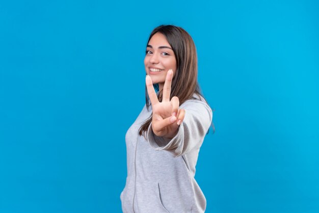 Giovane bella ragazza in grigio che guarda l'obbiettivo con il sorriso sul viso che fa il gesto di pace in piedi su sfondo blu