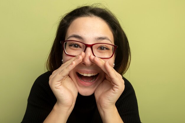 Giovane bella ragazza in un dolcevita nero e bicchieri gridando felice ed emozionato con le mani vicino alla bocca