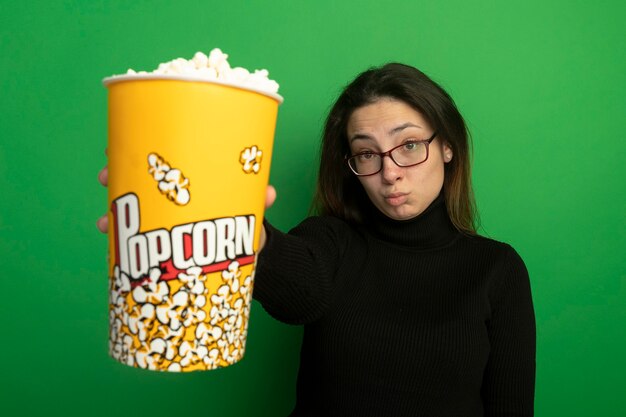 Giovane bella ragazza in un dolcevita nero e occhiali che mostrano secchio con popcorn confuso