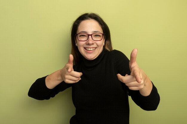 Giovane bella ragazza in un dolcevita nero e occhiali felice e positivo che punta con indice figners alla telecamera