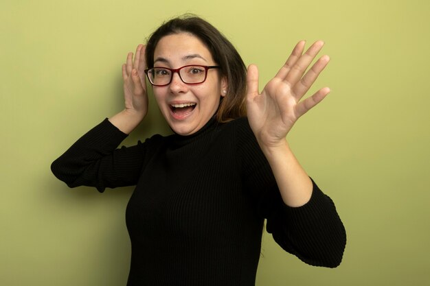 Giovane bella ragazza in un dolcevita nero e occhiali sorridendo allegramente con le mani alzate